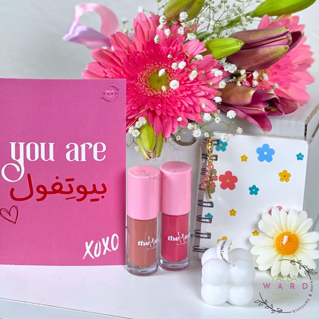 Two lipsticks, a pink card with flowers, and a small white bottle on a white surface.