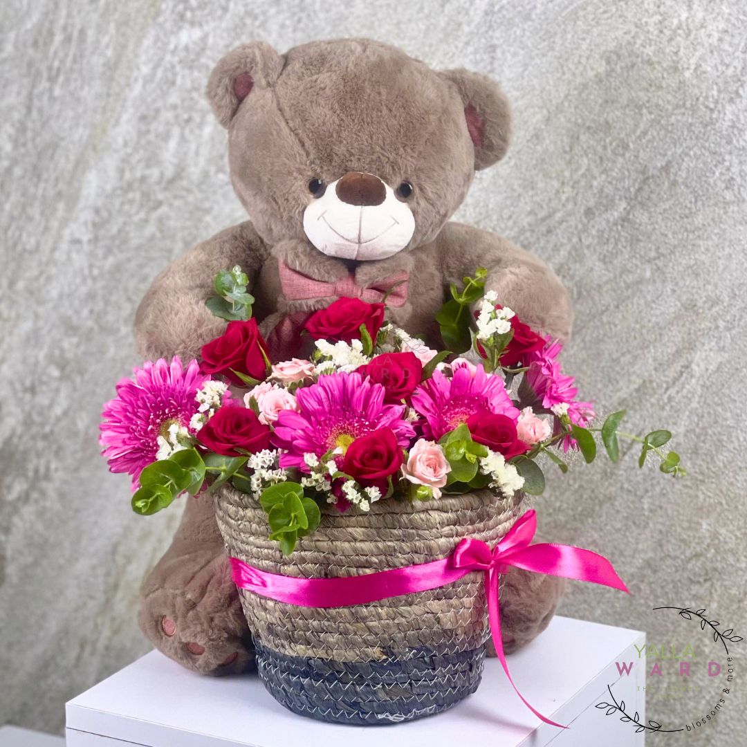 Teddy bear holding a basket of flowers with a pink ribbon against a gray background