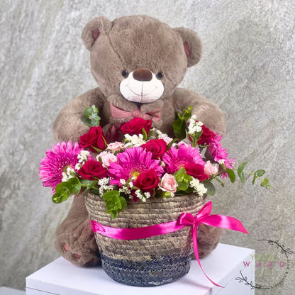 Teddy bear holding a basket of flowers with a pink ribbon against a gray background