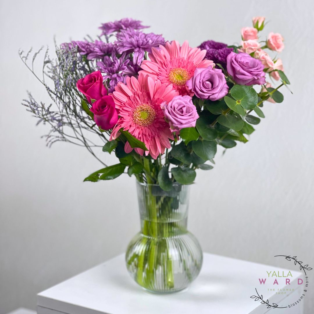 pink, purple, and green flowers in a clear vase on a white surface with 'Yalla Ward' branding.