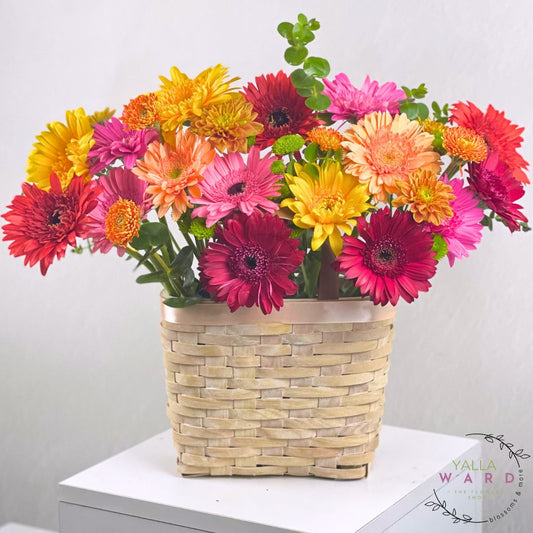 Colorful bouquet of flowers in a woven basket on a white surface with a light gray background.