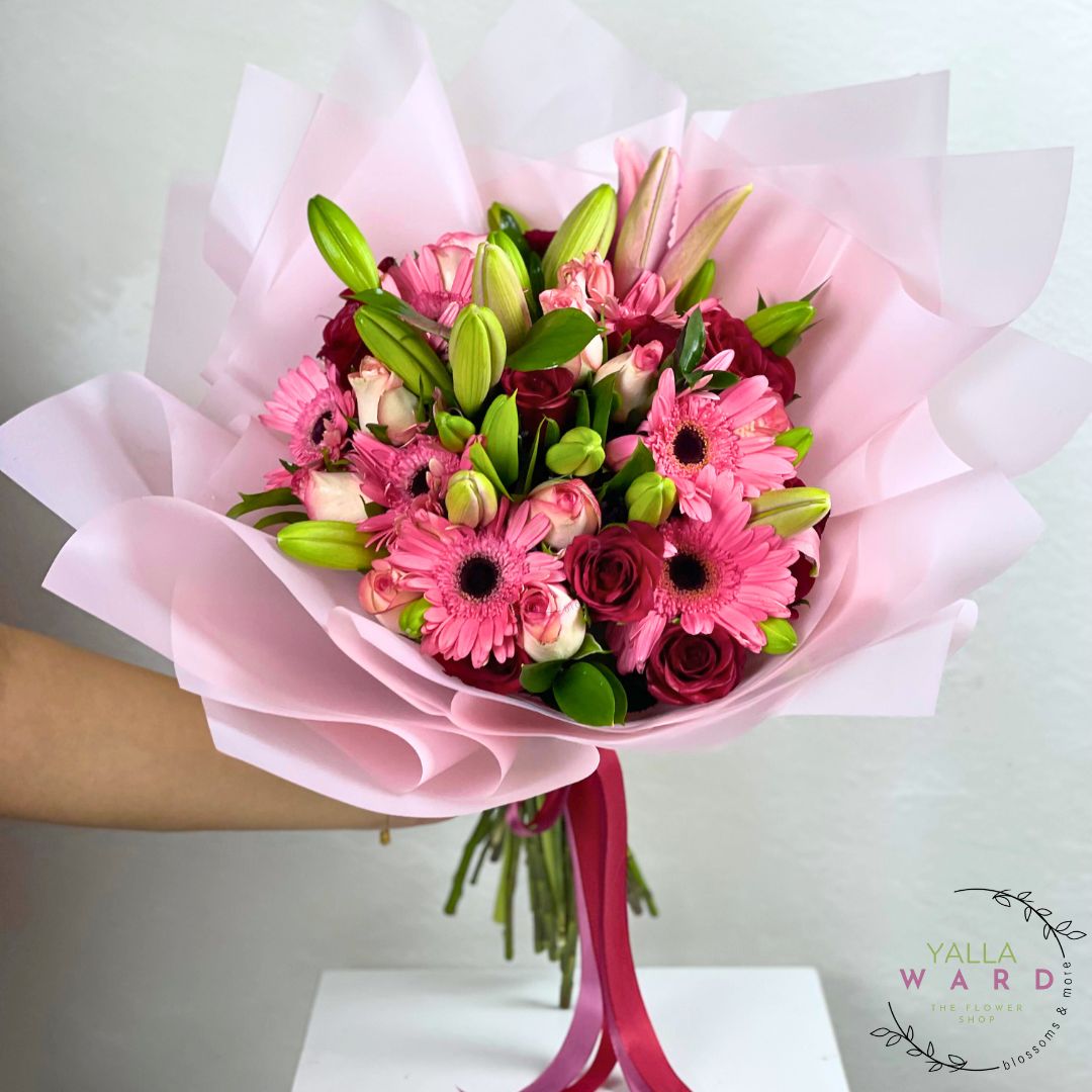 Bouquet of pink flowers with green leaves wrapped in pink paper, held by a person.