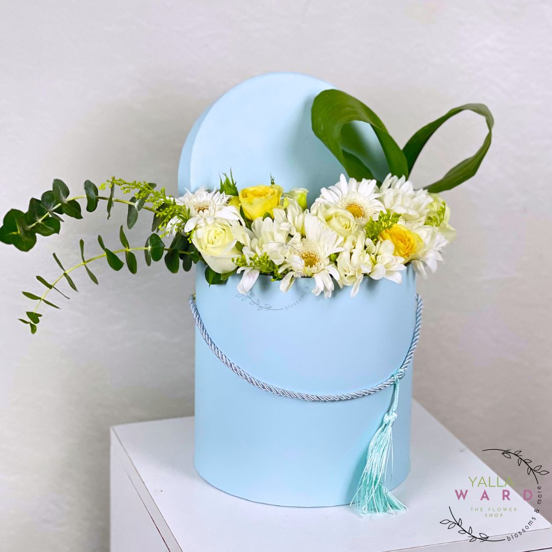 Floral arrangement in a light blue box with greenery on a white surface.
