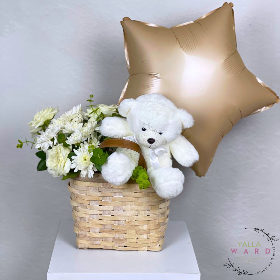 White teddy bear, basket of flowers, and gold star balloon on a white surface with 'Yalla Ward' branding.