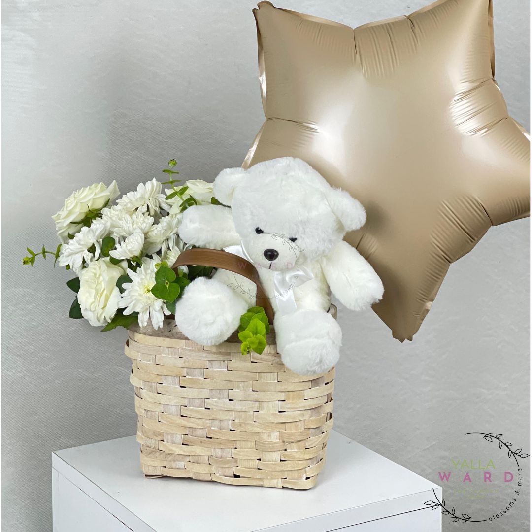 White teddy bear and flowers in a basket with a gold star balloon on a white surface.