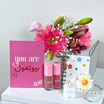 Pink card with 'you are beautiful' text, two lipsticks, flowers, and a candle on a white surface.
