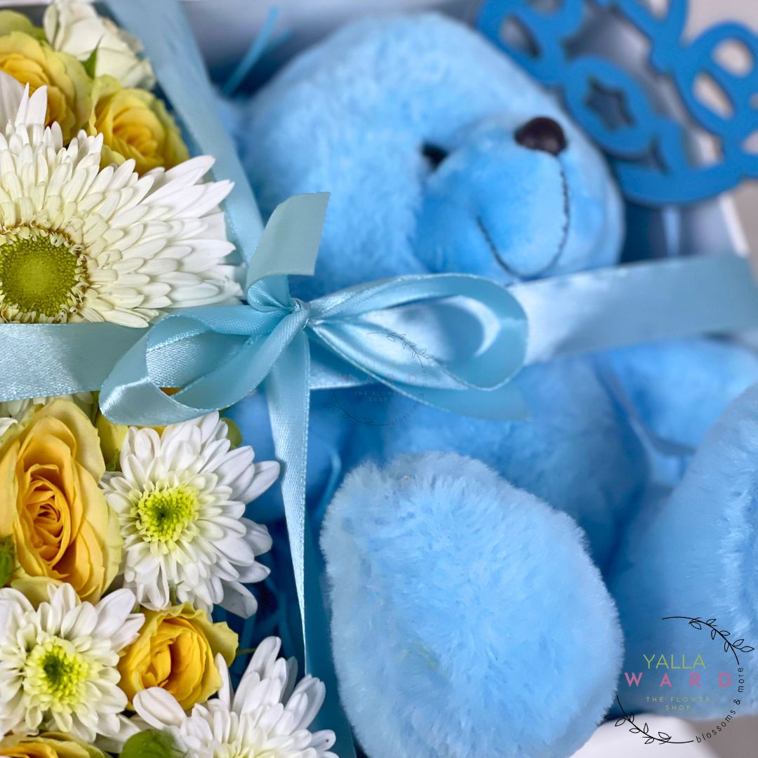 Blue teddy bear with a bouquet of flowers and a decorative card on a white background.