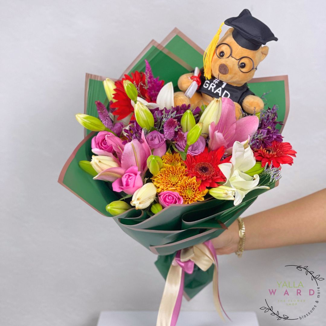 Colorful bouquet with a teddy bear wearing a graduation cap and 'GRAD' shirt, held by a person.