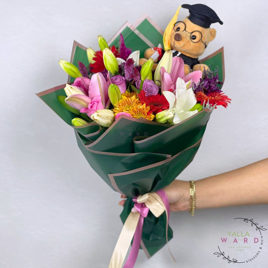 Bouquet of flowers with a teddy bear wearing a graduation cap, held by a person against a white background.