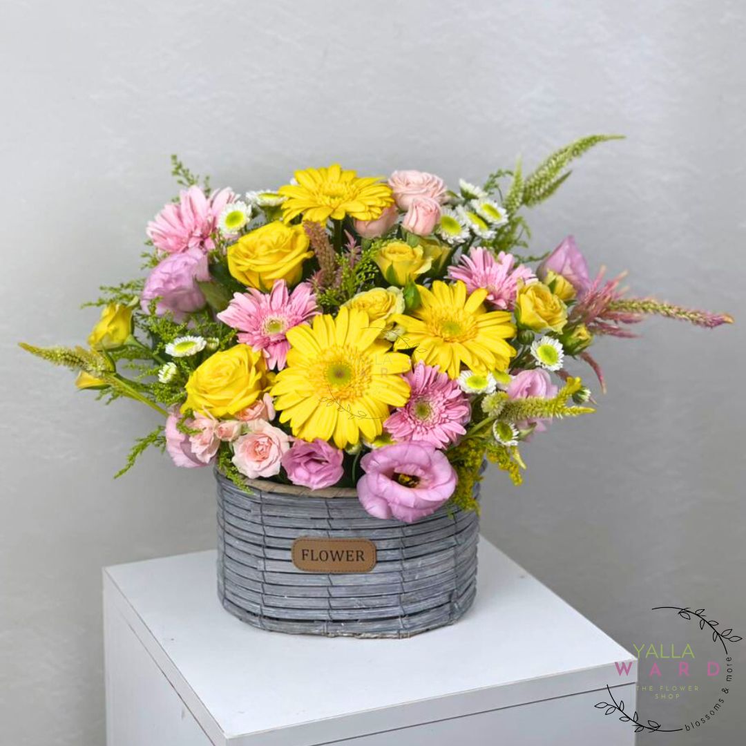 Colorful flower arrangement in a gray basket on a white surface with a light gray background.