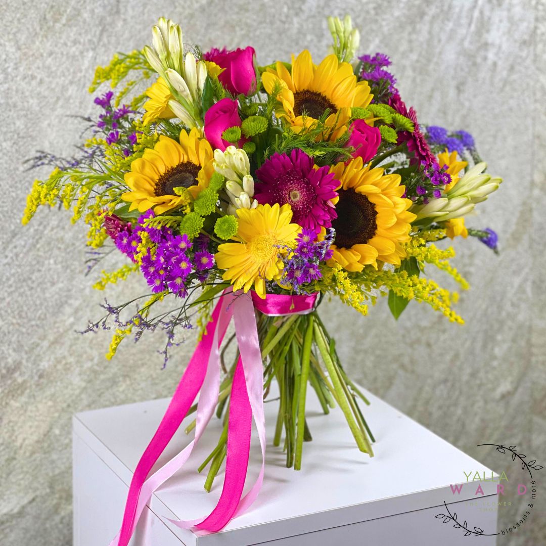 Bouquet of flowers with sunflowers, pink and purple flowers, and a pink ribbon on a white surface.
