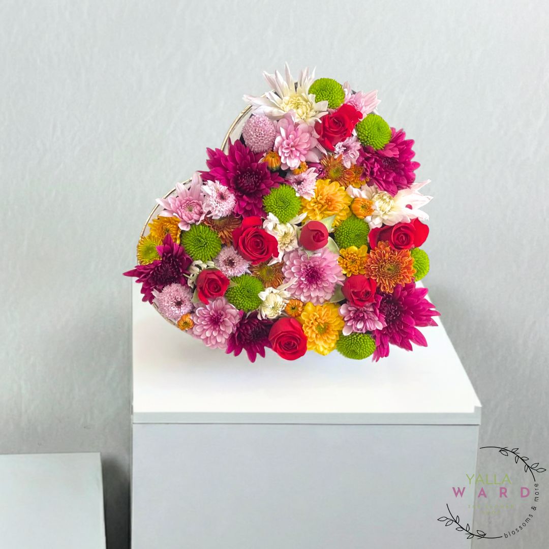 Heart-shaped flower arrangement with colorful flowers on a white surface against a light gray background.