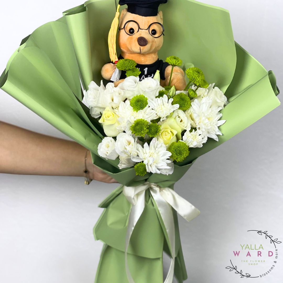 Bouquet of white flowers with a teddy bear wearing a graduation cap, held by a person against a white background. Brand 'Yalla Ward' visible.